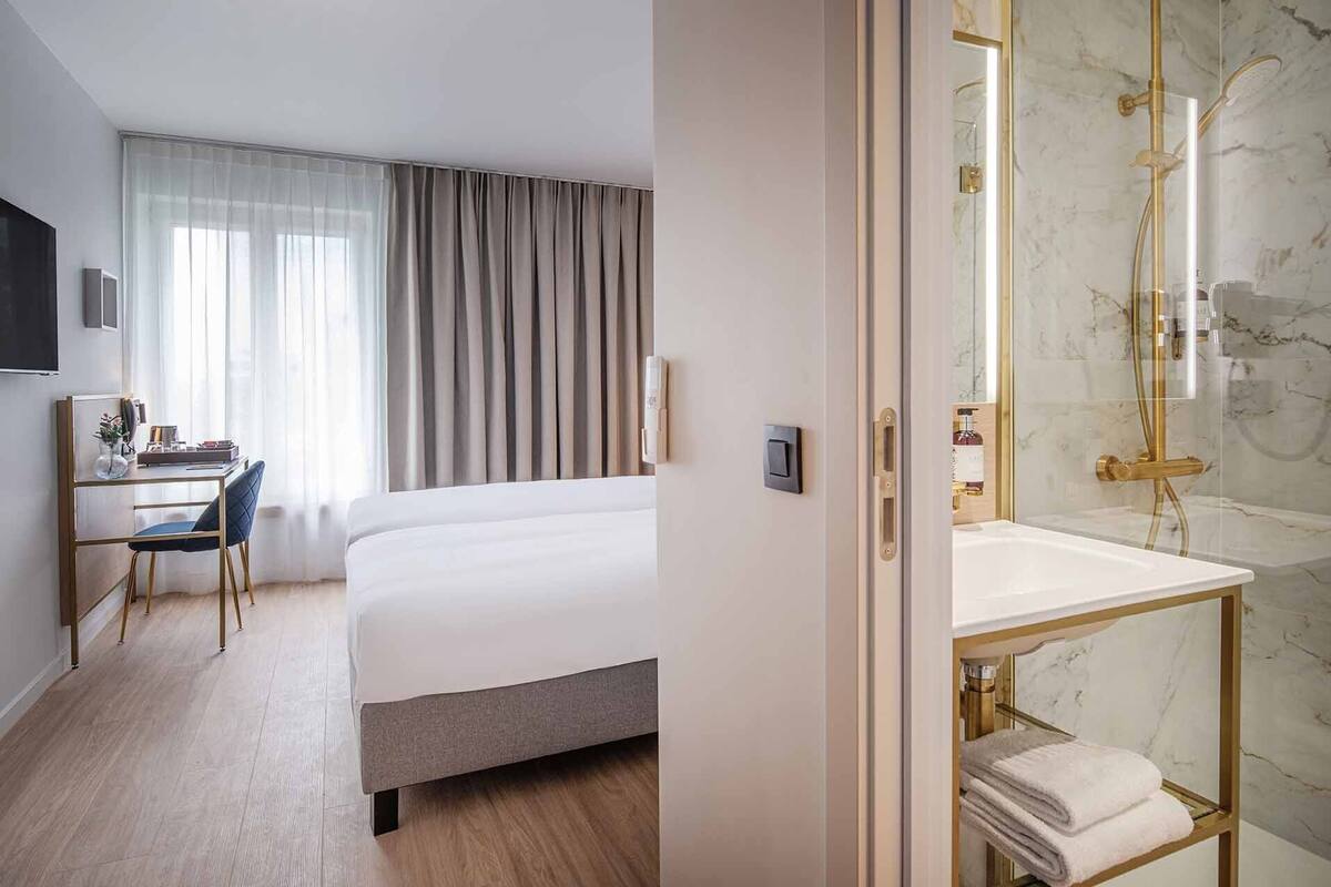 A hotel room with a bed, a desk with a chair, a shower with a marble wall, and a bathroom with a sink and towel rack.