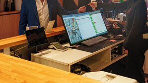 A reception desk with a computer displaying a schedule, a phone, and a tablet. There are framed pictures on the wall and a plant in the background. - Jill Hotel Brussels (Brussels)