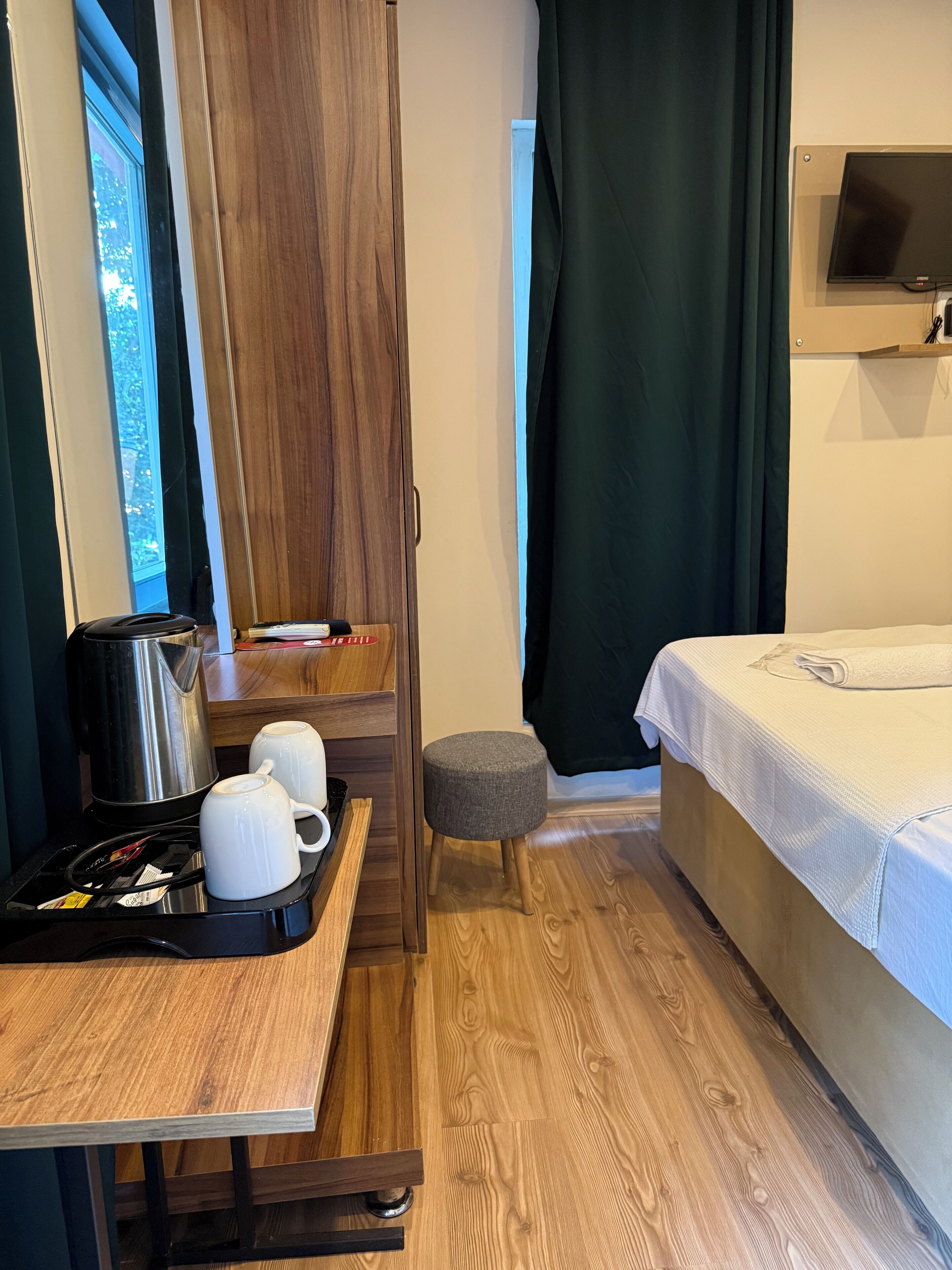Habitación compacta con piso de madera, una cama con cubrecama blanco, un taburete pequeño, una mesa con tetera y tazas, y un televisor montado en la pared.