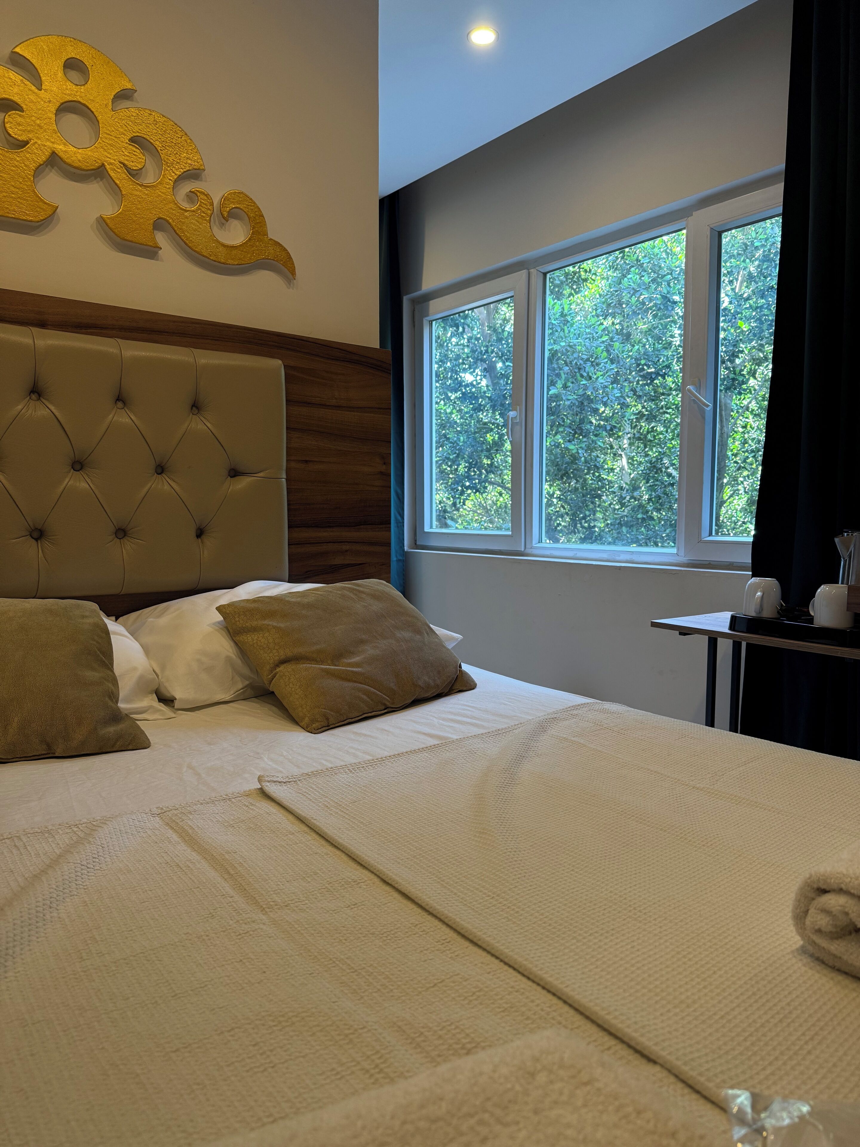 A neatly made bed with a tufted headboard, a window with a view of greenery, and a small table with a lamp.