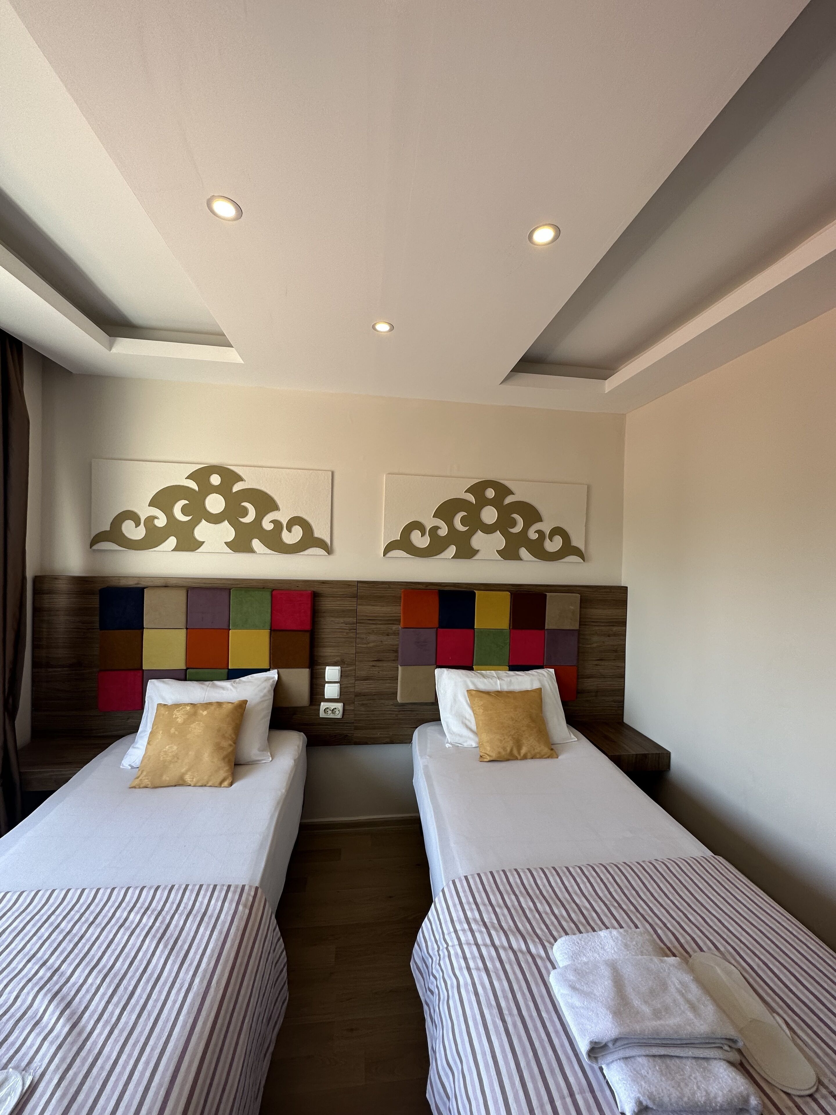 A hotel room with two beds, a wooden headboard, a colorful wall panel, and a ceiling with recessed lighting.