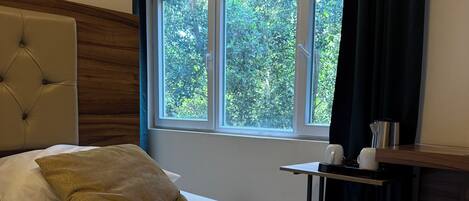 Habitación de hotel con una cama grande, cabecera de madera, ventana con vistas a árboles y una mesita con una tetera y tazas.