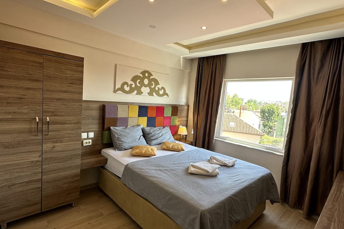 A modern bedroom with a wooden wardrobe, a bed with a grey bedspread, colorful pillows, and a window with curtains.