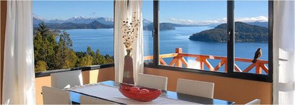 Un comedor con una mesa y sillas, un tazón rojo y vistas a un lago con montañas al fondo.