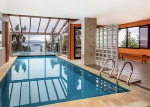 Una piscina interior moderna con paredes de vidrio transparente, vistas al mar y una zona de terraza de madera.