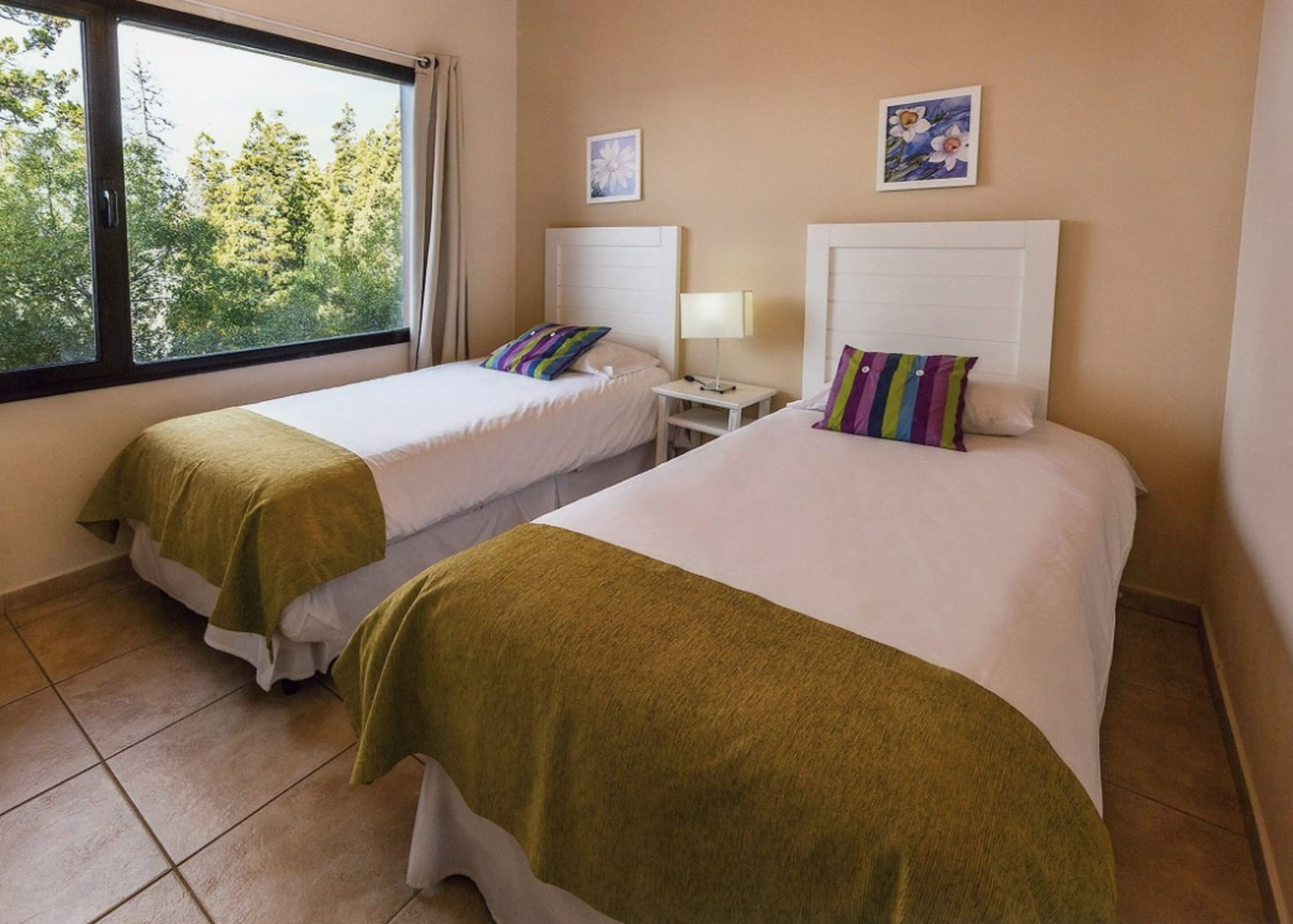 A hotel room with two beds, a window with a view of trees, and a small bedside table with a lamp.