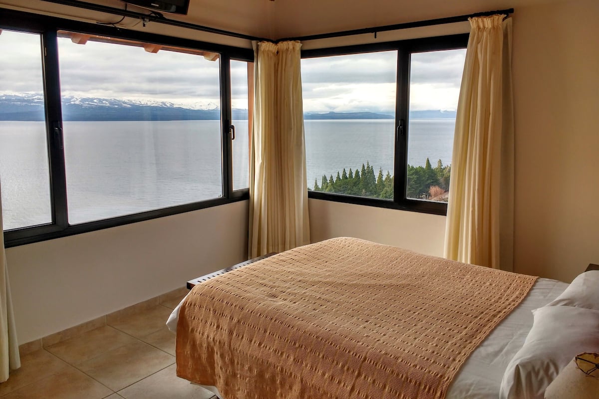 A bedroom with a bed, large windows, and a view of a lake and mountains.