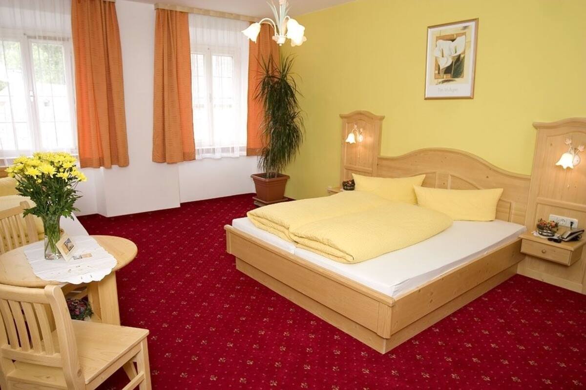 Ein Hotelzimmer mit einem hölzernen Bett, einem roten Teppich, einer Topfpflanze, einem Tisch mit Stühlen und einem fenster mit Vorhängen.