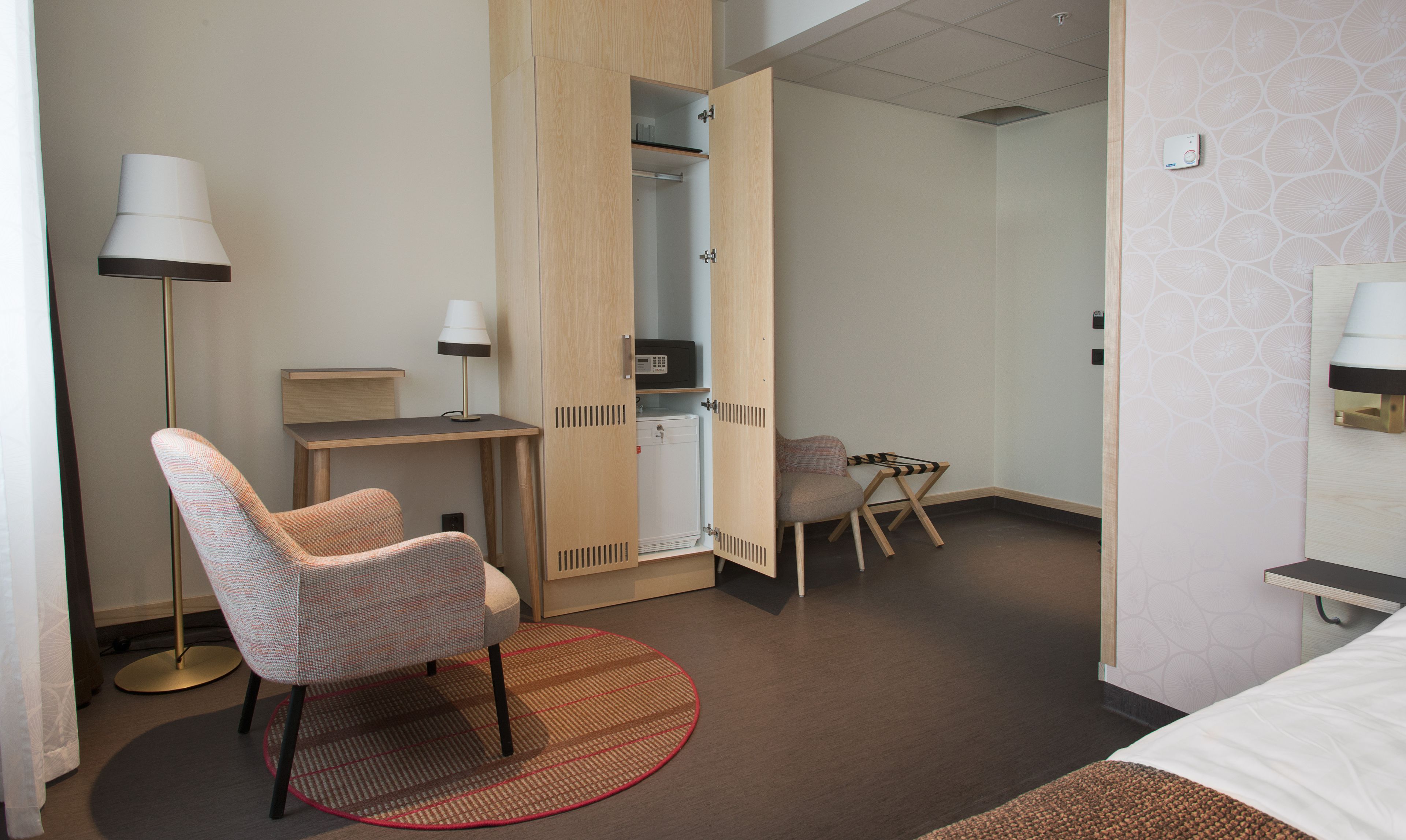 Una habitación de hotel con cama, escritorio, silla, armario y una mesita de noche con lámpara.