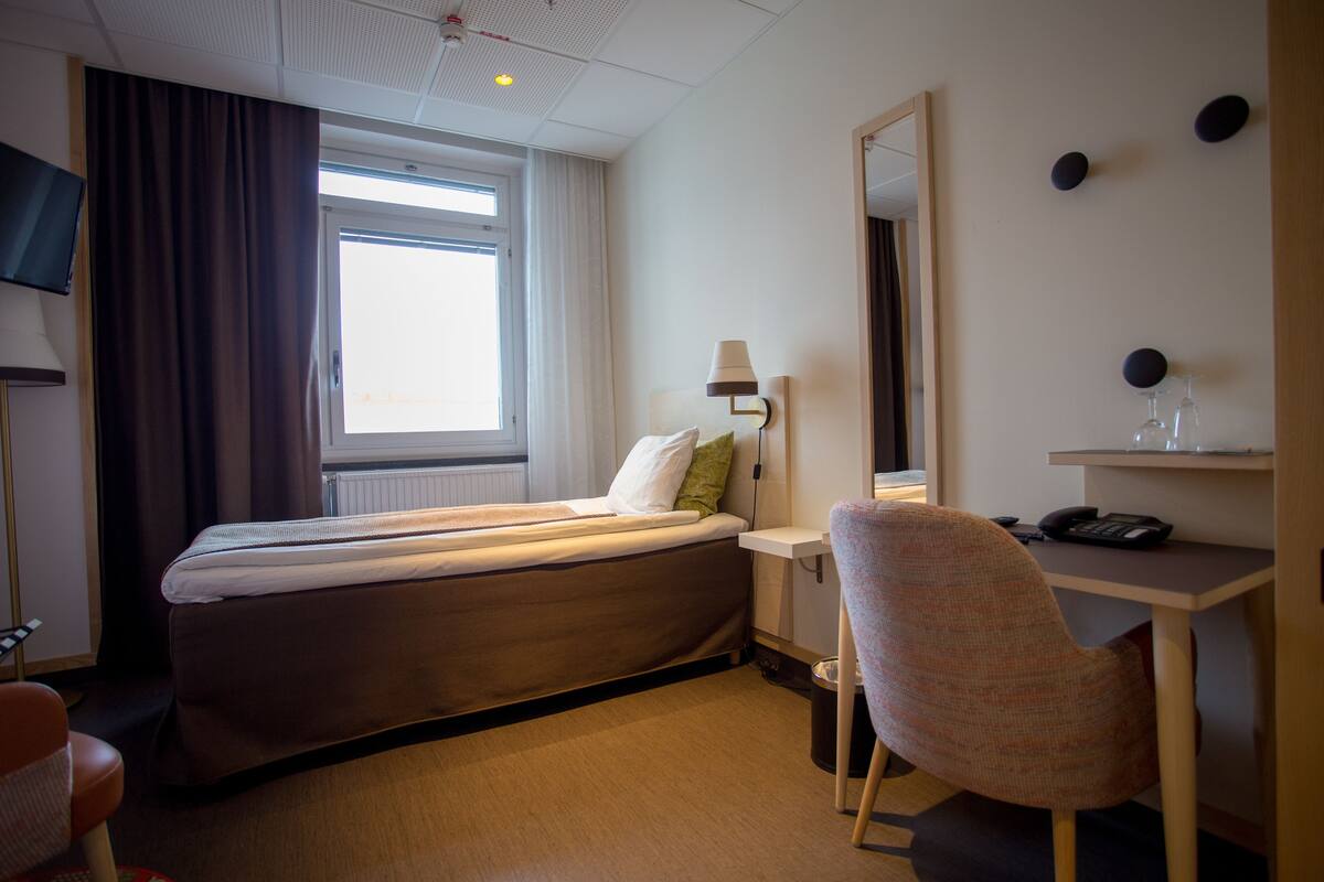 A hotel room with a bed, a desk with a lamp, a chair, a TV, and a window with curtains.