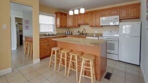 A kitchen with wooden cabinets, a white refrigerator, a microwave, and a sink. There are three wooden stools at the island. - Ocean Lodge Hotel South (Ocean City)