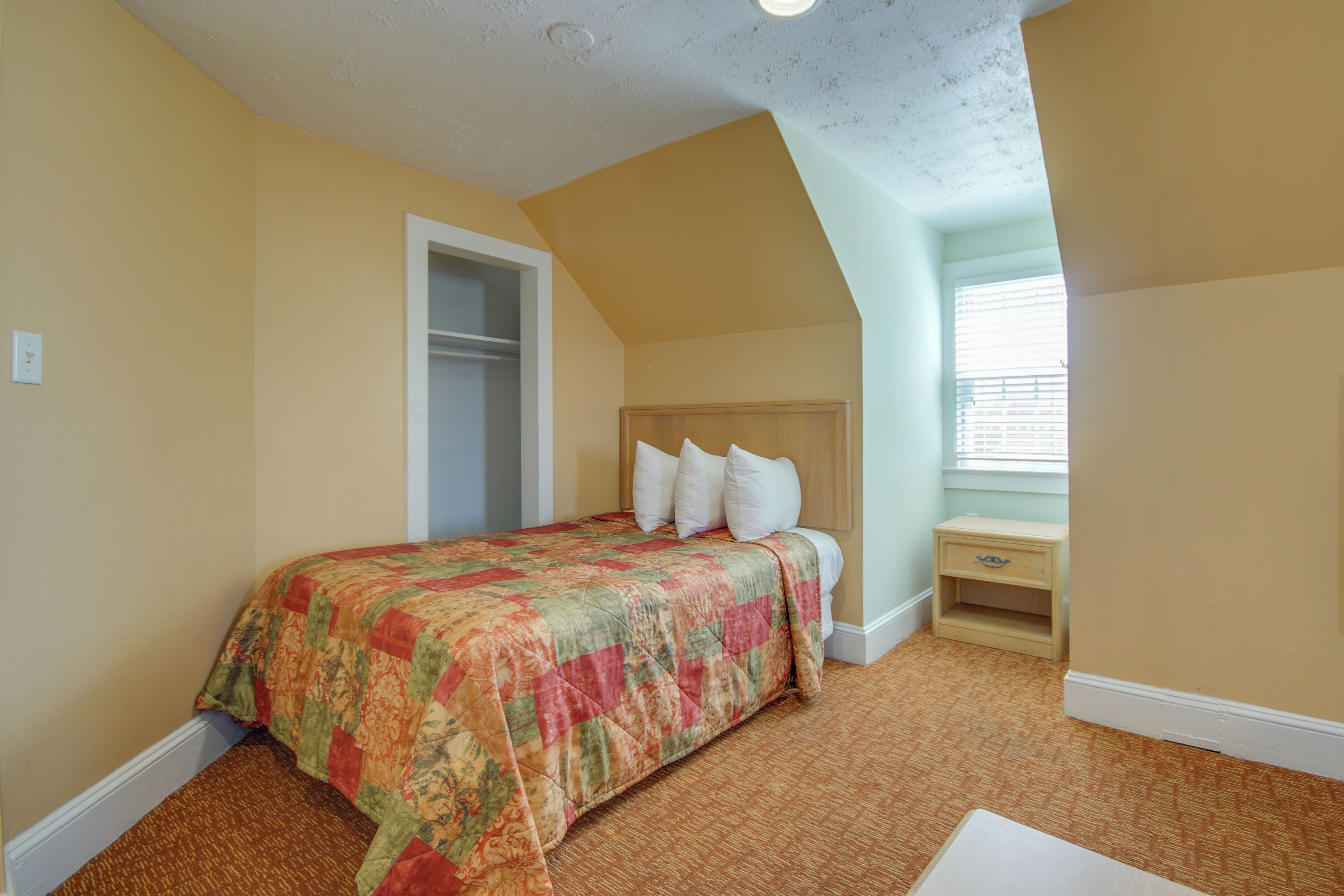 Une chambre à coucher comprenant un lit, une table de chevet, une fenêtre avec des stores et une porte donnant sur une autre pièce.