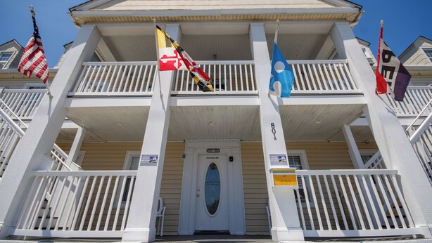 A two-story building with a white facade, a balcony on the second floor, and multiple flags including the American flag. - Ocean Lodge Hotel South (Ocean City)