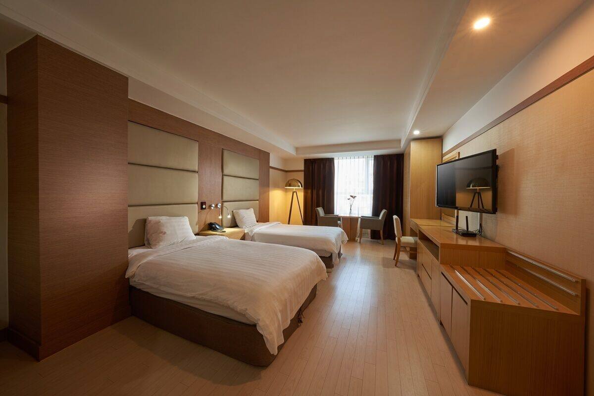 A hotel room with two beds, a desk, a chair, a TV, and a lamp.
