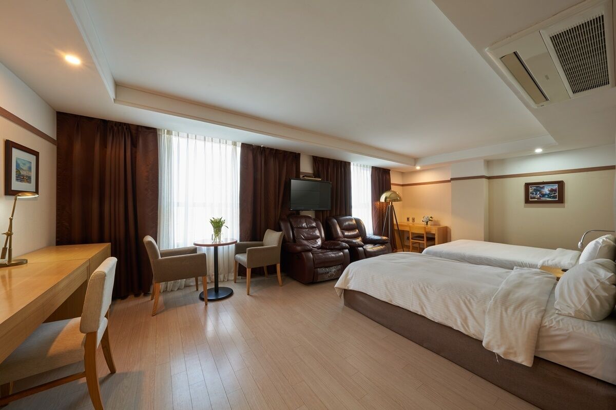 A hotel room with a bed, desk, chairs, a TV, and a small table with a lamp.