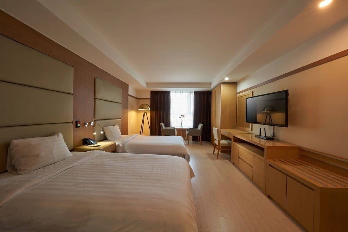 A hotel room with two beds, a desk, a TV, and a window with curtains.