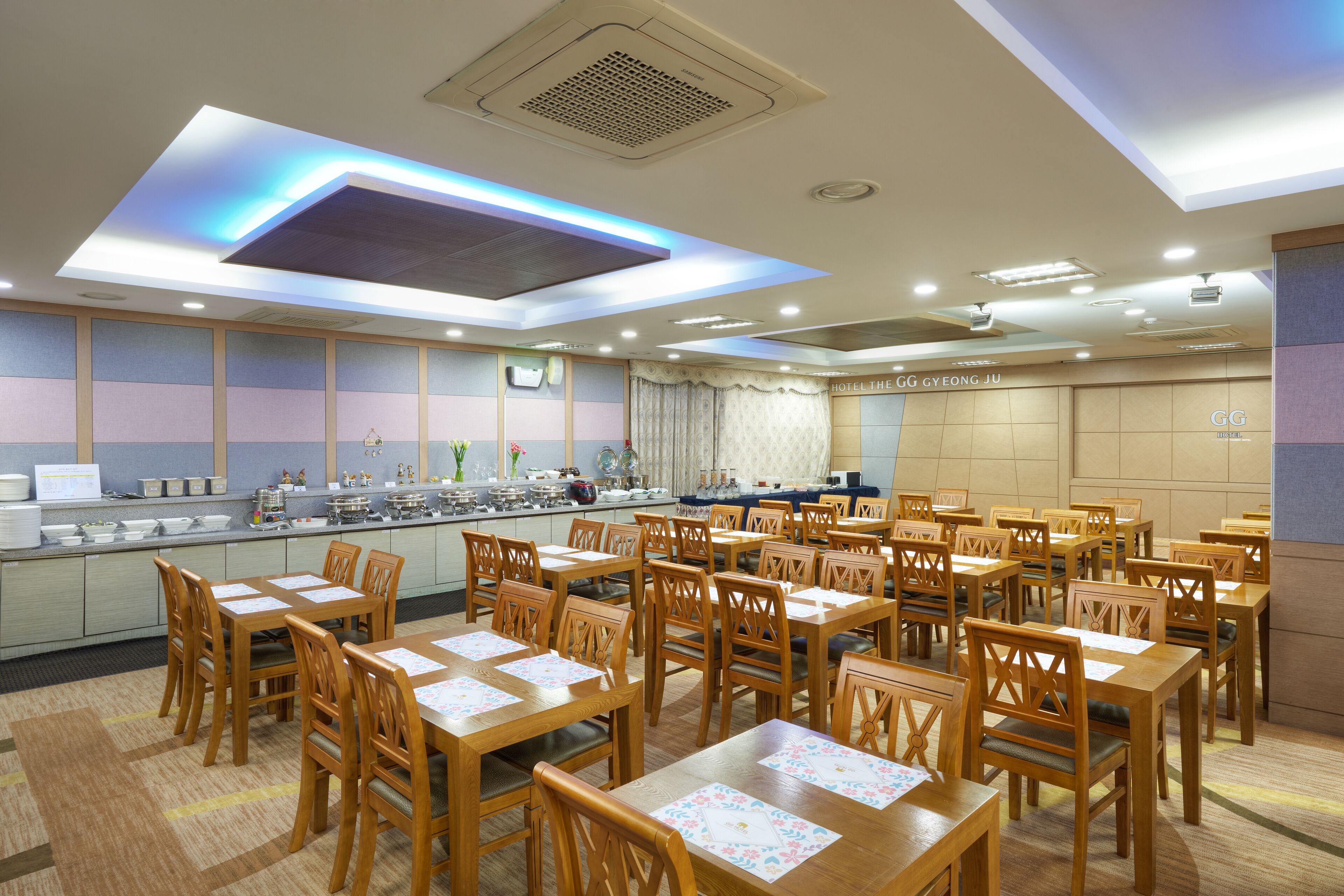 Une salle à manger d’hôtel avec de nombreuses tables et chaises en bois, un buffet et un plafond doté d’un éclairage encastré.