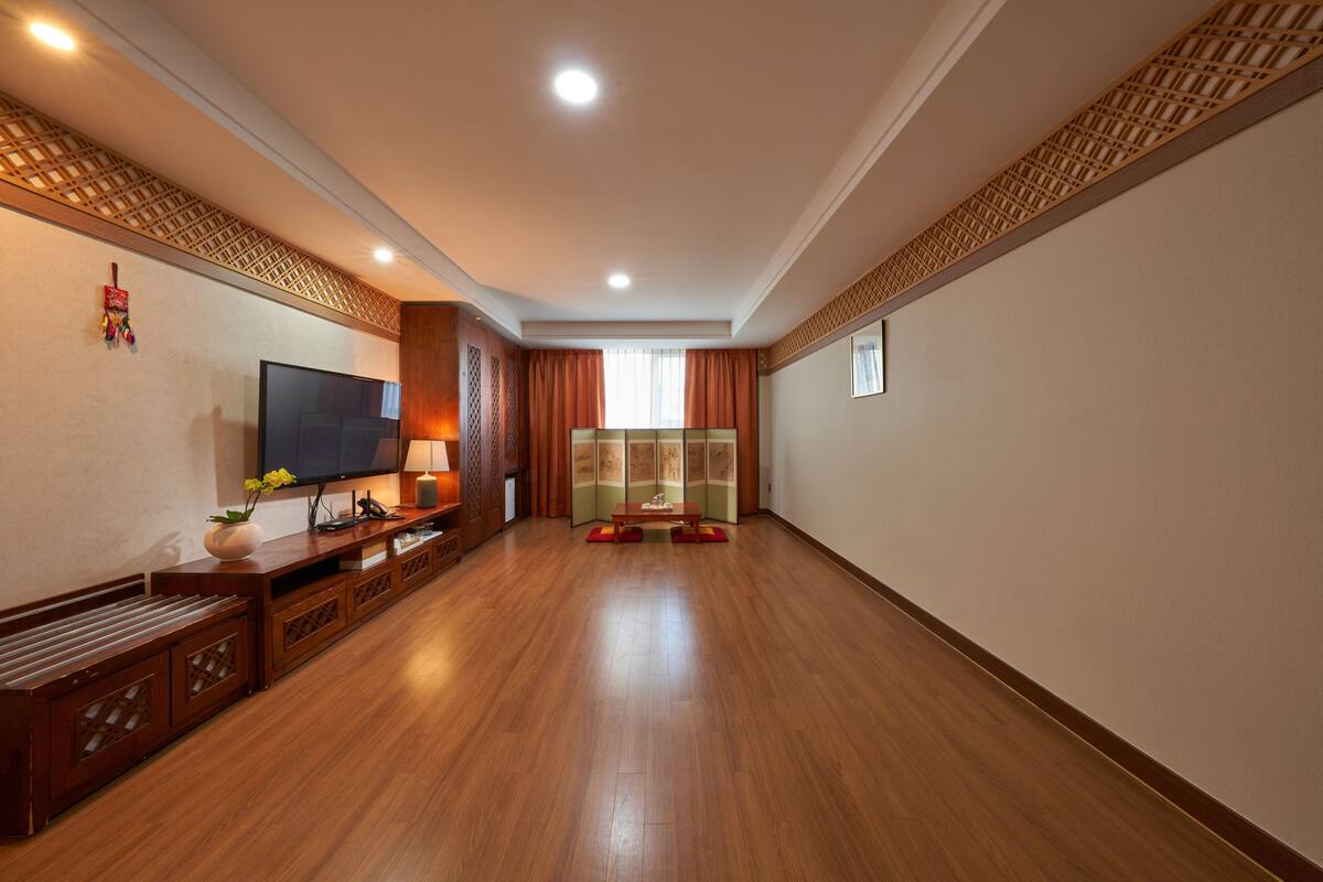 A spacious room with a wooden floor, a flat-screen TV, a wooden entertainment unit, and a traditional-style window with curtains.