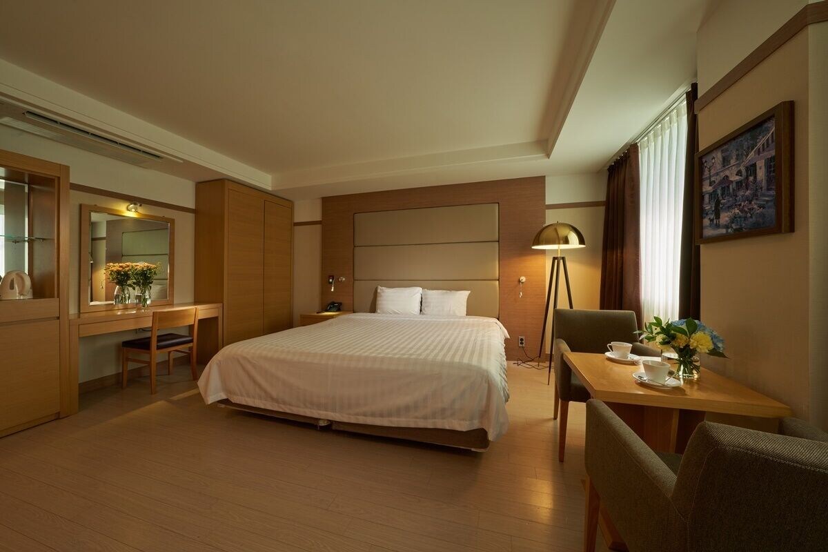 Une chambre d’hôtel avec un grand lit, un bureau avec une chaise, une petite table avec une chaise, une lampe et un tableau au mur.