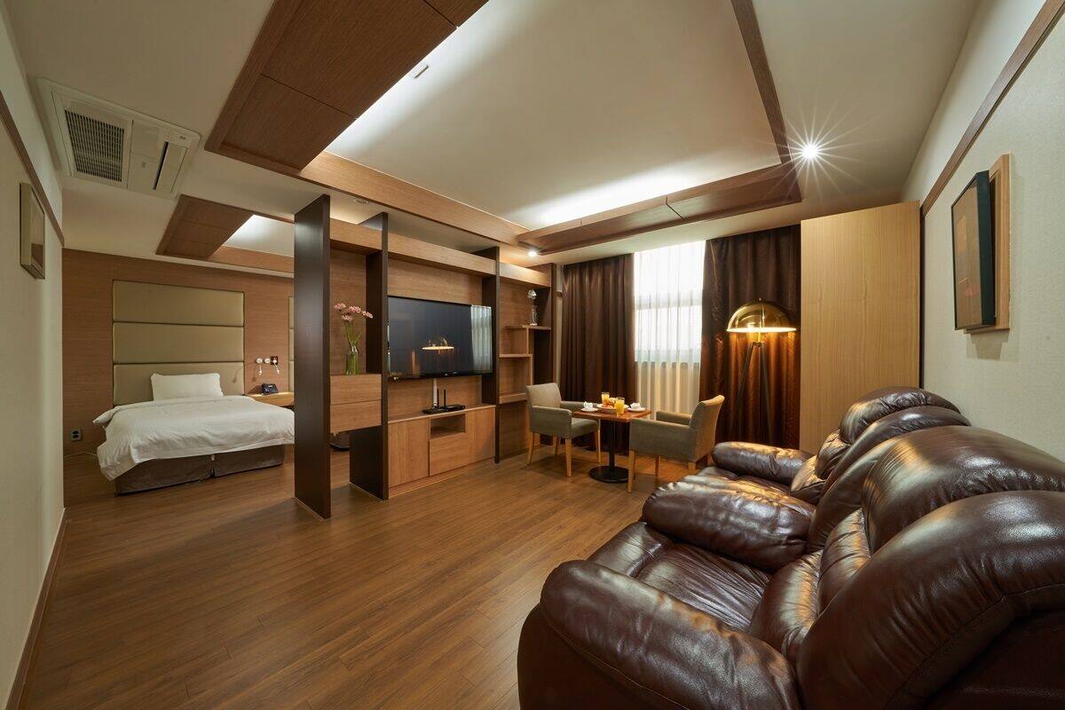 A modern hotel room with a wooden floor, a leather sofa, a bed, a TV, a desk, and a dining area.