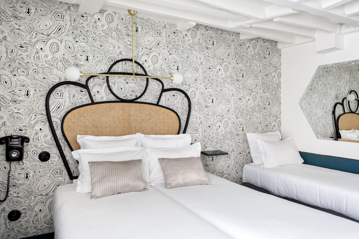 Et hotelværelse med tapet med mønster, en fletningssenggavl, hvide sengetøj og et natbord med lampe.