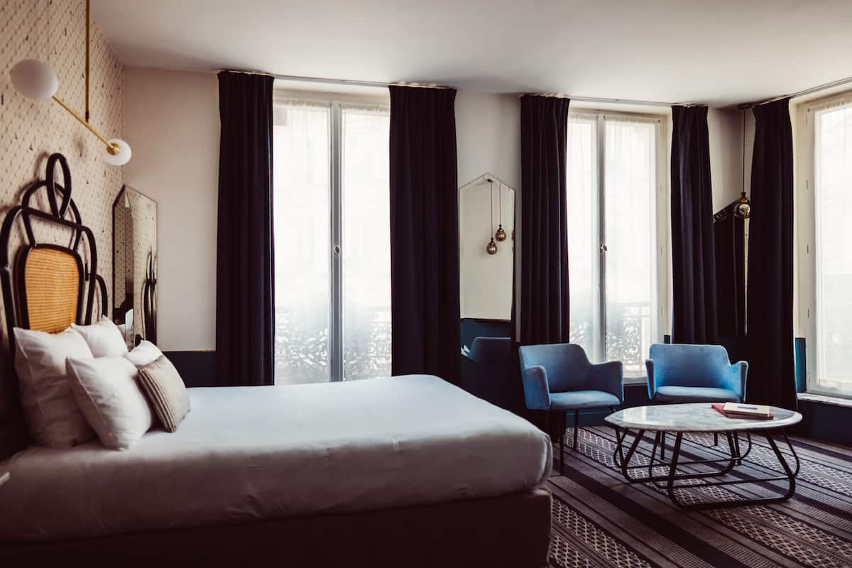 Et hotelværelse med en seng, to blå lænestole, et rundt sofabord og store vinduer med mørke gardiner.