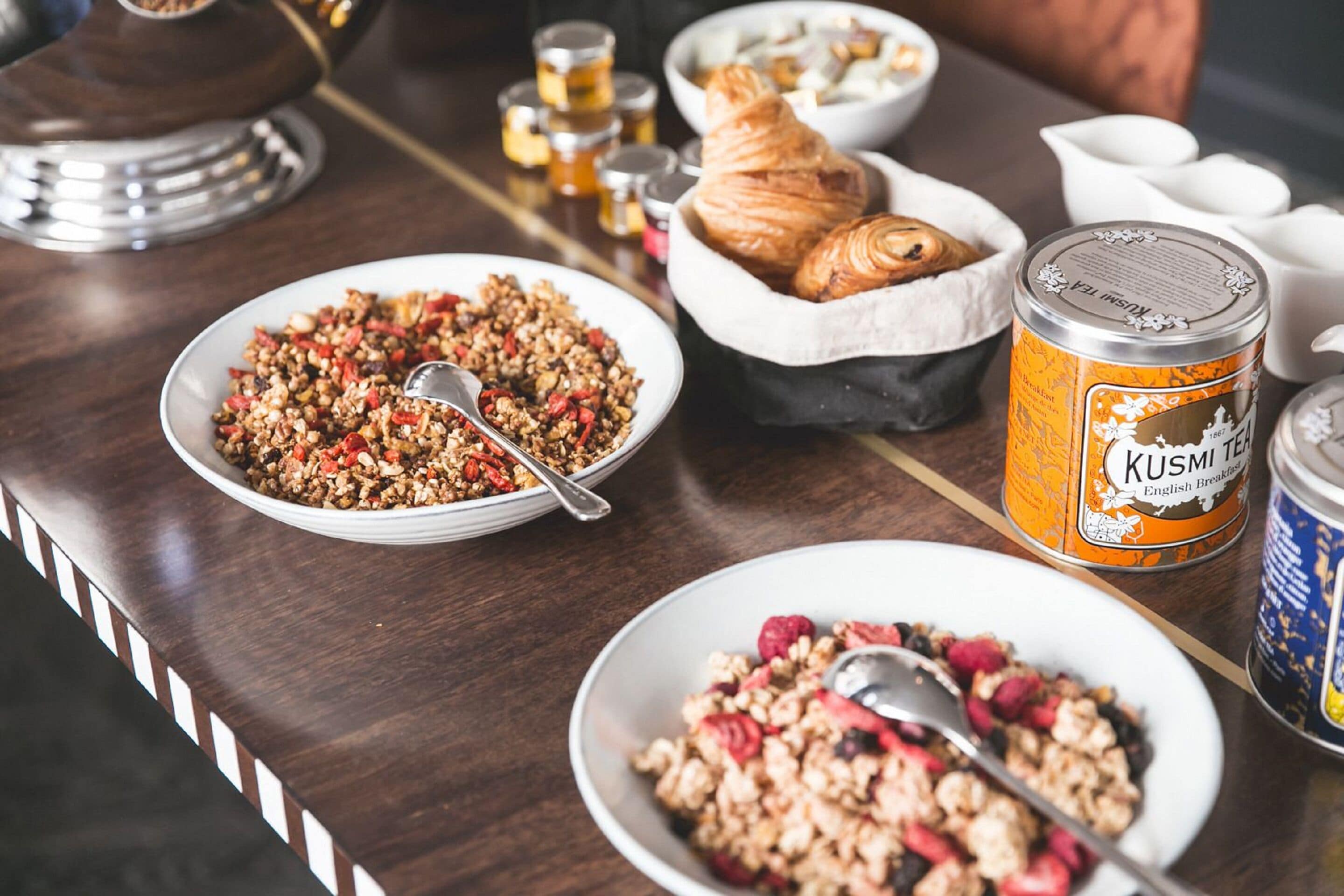 A breakfast table with bowls of cereal, a croissant basket, and a can of tea.