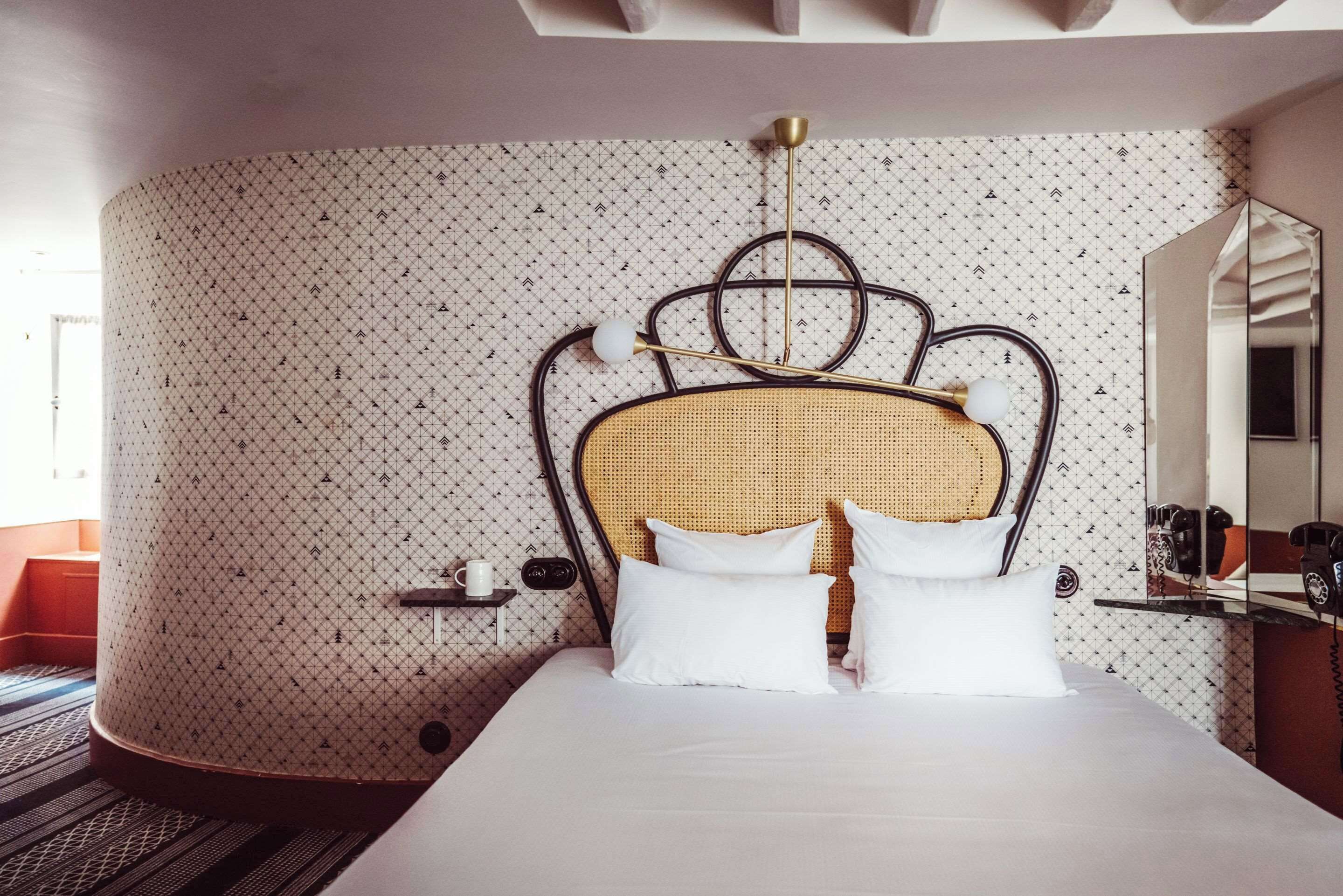 Une chambre d’hôtel avec un lit, une tête de lit ornée d’un motif tissé, une table de chevet avec une lampe et un mur décoré d’un motif.