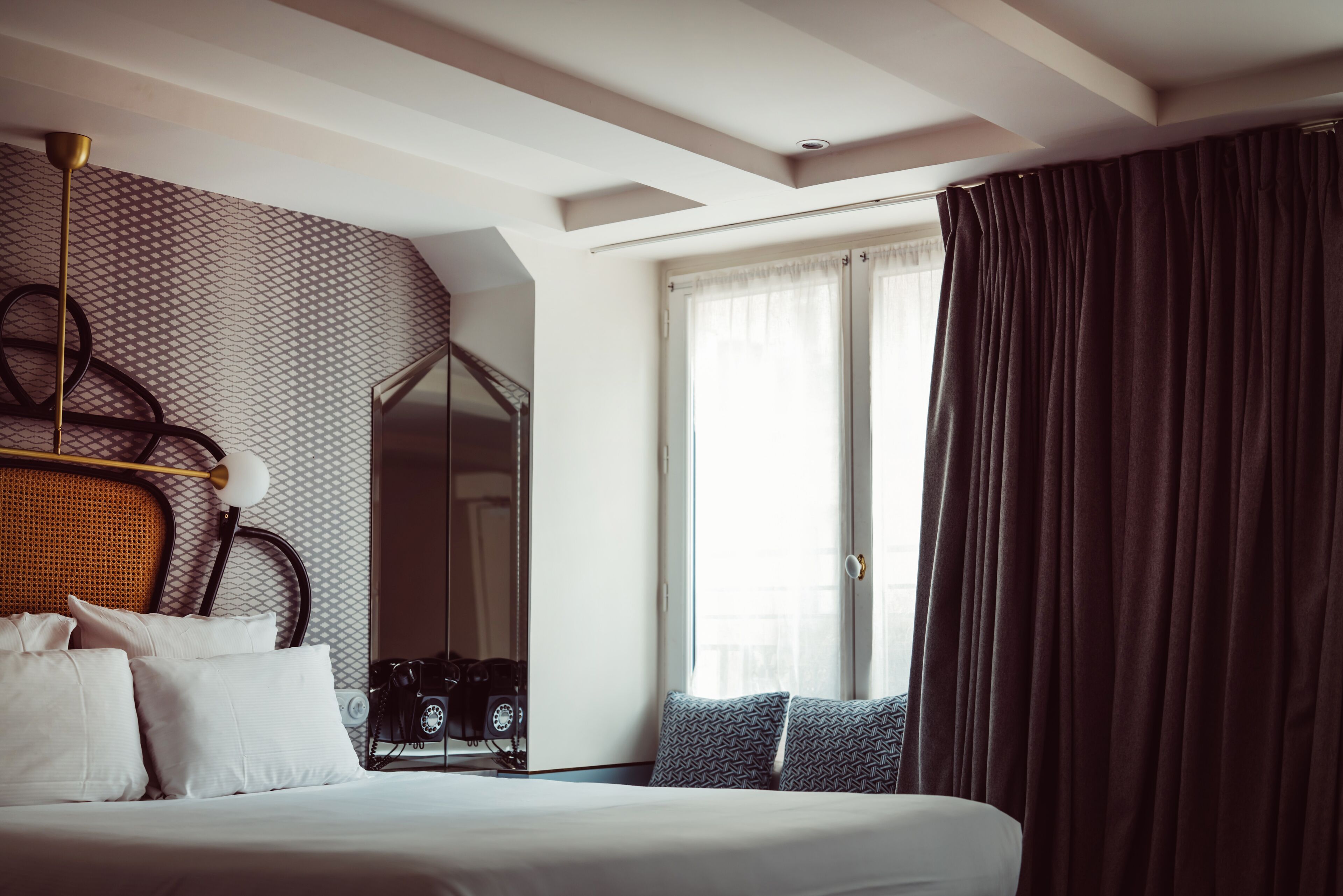 Een hotelkamer met een bed, een grote spiegel, een camera op het nachtkastje en een raam met gordijnen.