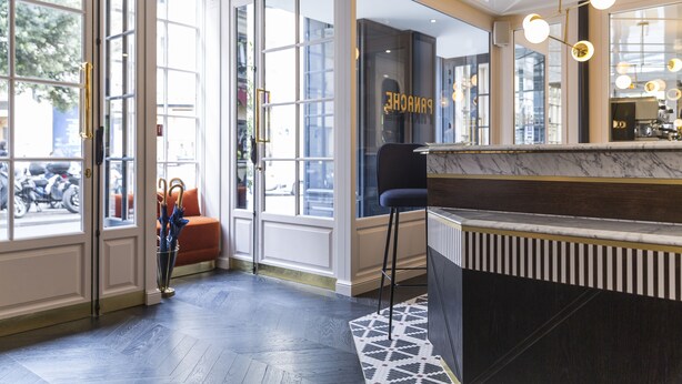 A hotel lobby with a reception desk, a patterned rug, and a view of the street through large glass doors. - Hotel Panache (Paris)