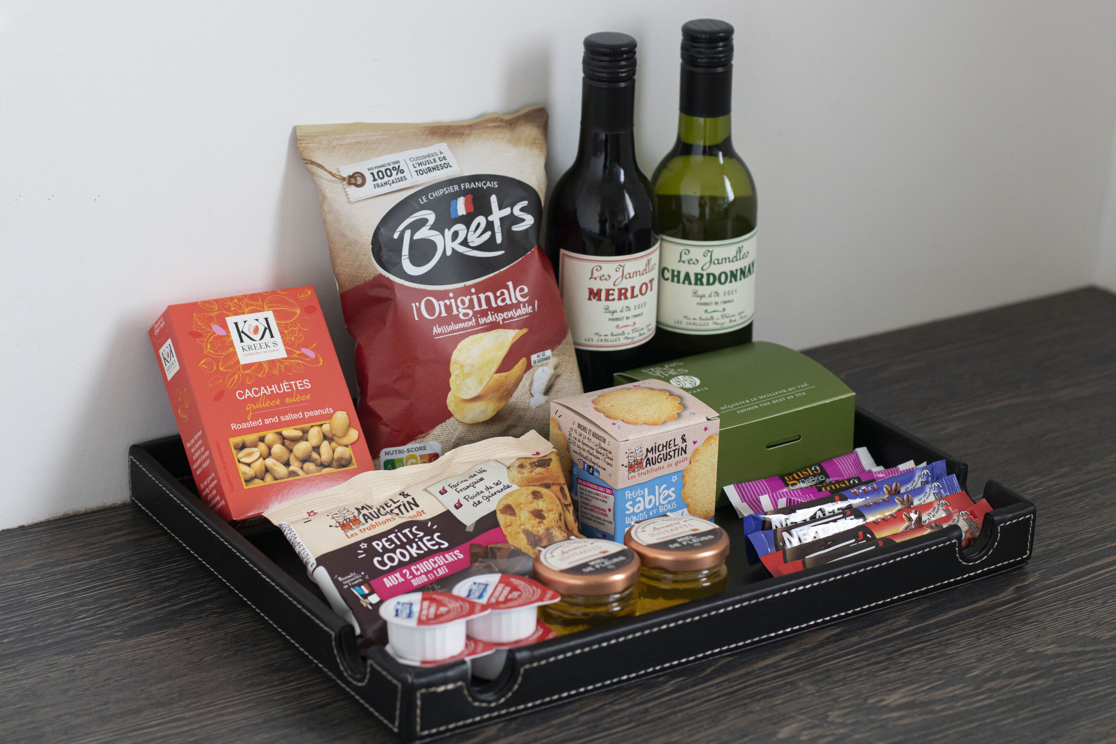 En bakke med forskelligt mad, heriblandt en pose Brets, en flaske Merlot og en kasse Les Jambelots Chardonnays.
