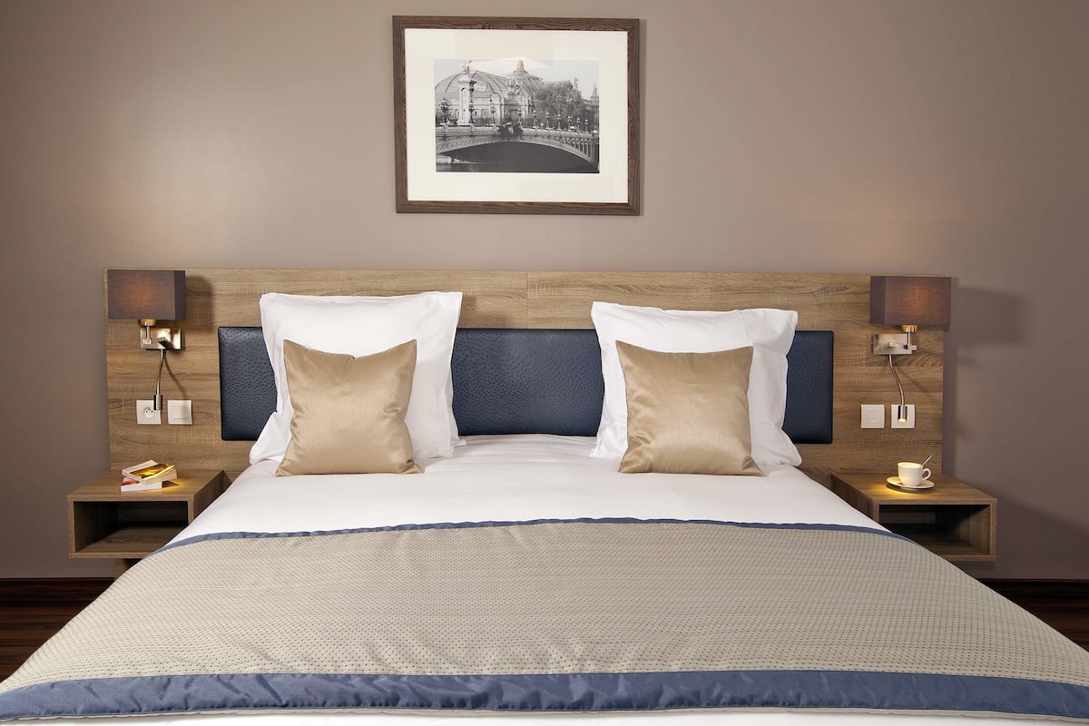 A hotel room with a large bed, two bedside tables with lamps, a framed picture on the wall, and a small table with a cup and saucer.