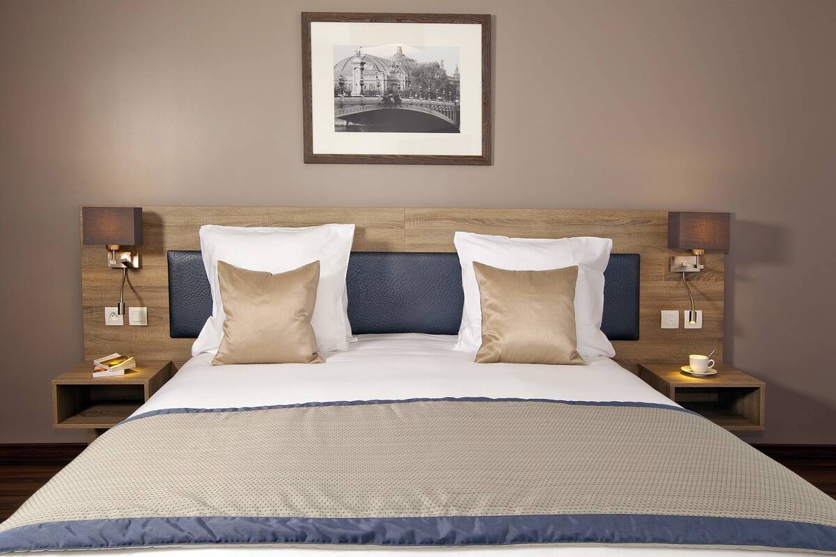 Een hotelkamer met een groot bed, twee nachtkastjes met lampen, een ingelijst schilderij aan de muur en een klein tafeltje met kop en schotel.