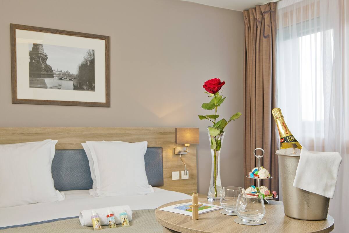 Een hotelkamer met een bed, een nachtkastje met een vaas met een rode roos, een fles champagne en een klein dessertplateau.