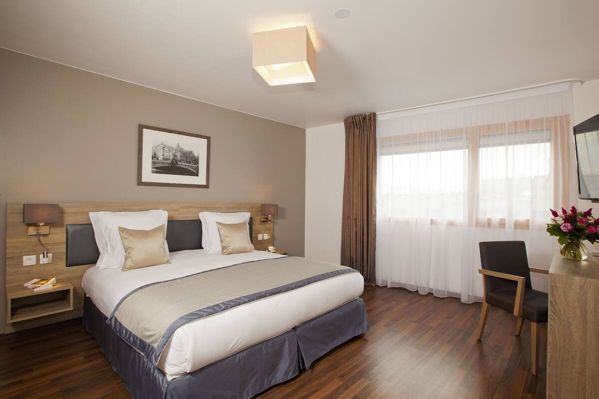 Hotelkamer met een groot bed, nachtkastjes, een bureau, een stoel, een lijst met een afbeelding aan de muur en een raam met gordijnen.