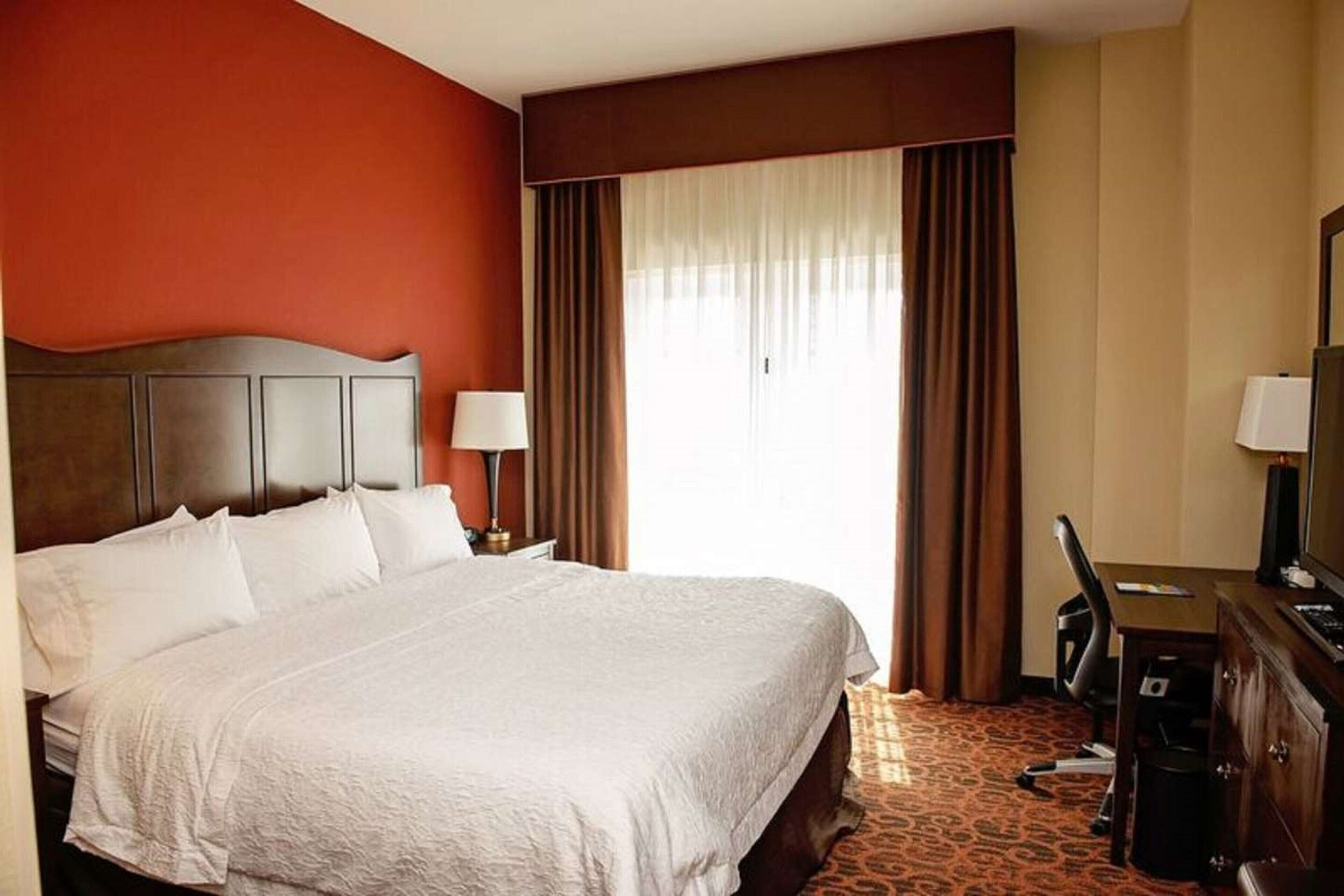Ein Hotelzimmer mit einem großen Bett, einem Schreibtisch mit Computer, einem Fernseher und einem Fenster mit Vorhängen.