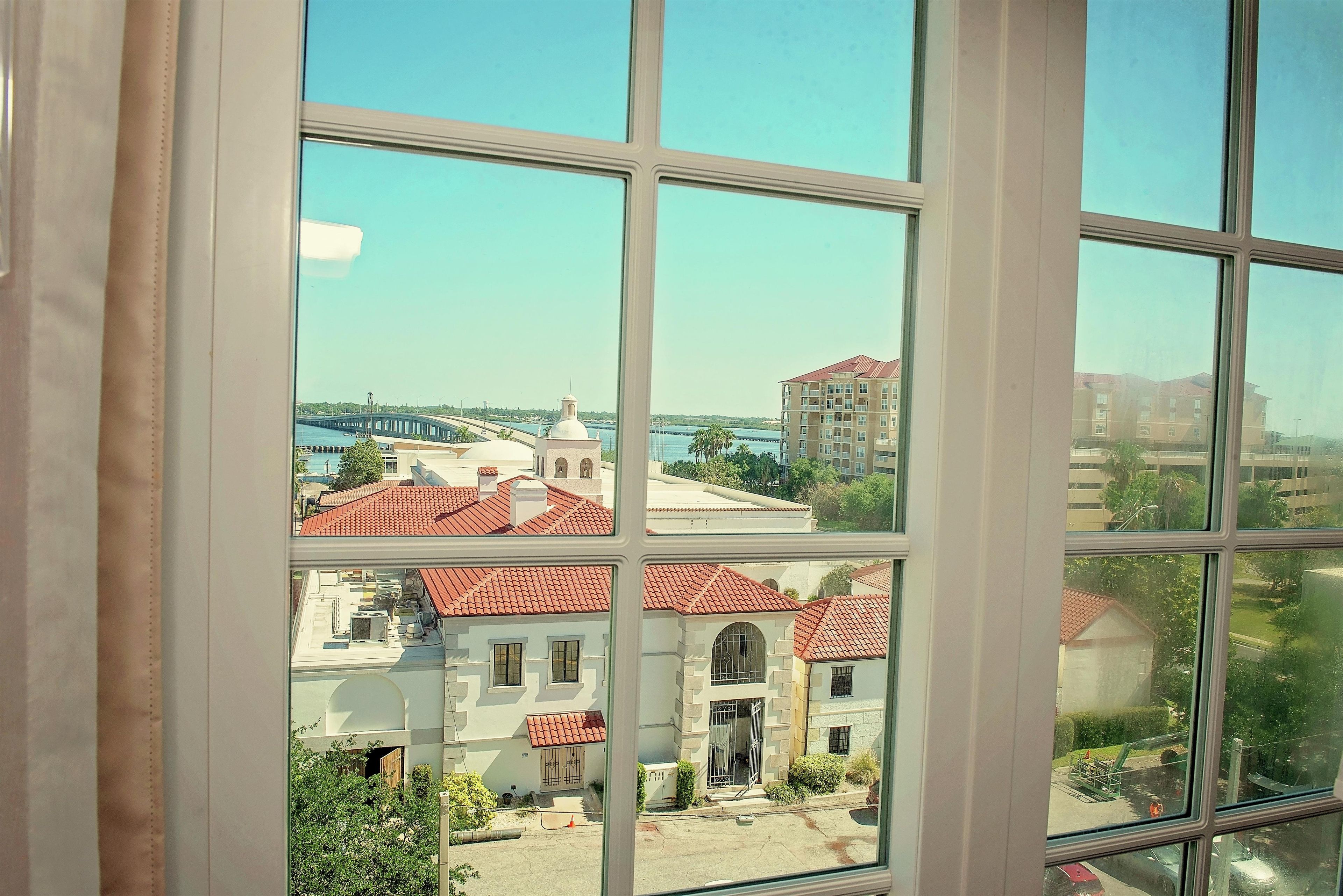 A view through a window of a building with a red-tiled roof, a white facade, and a clear blue sky.