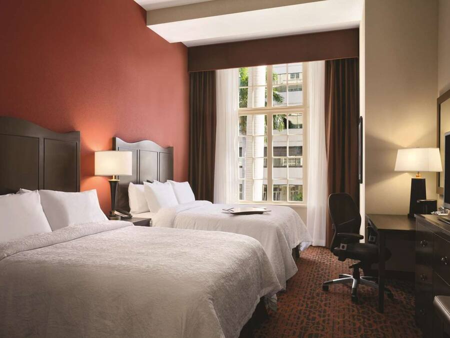 A hotel room with two beds, a desk, a chair, a TV, and a window with curtains.