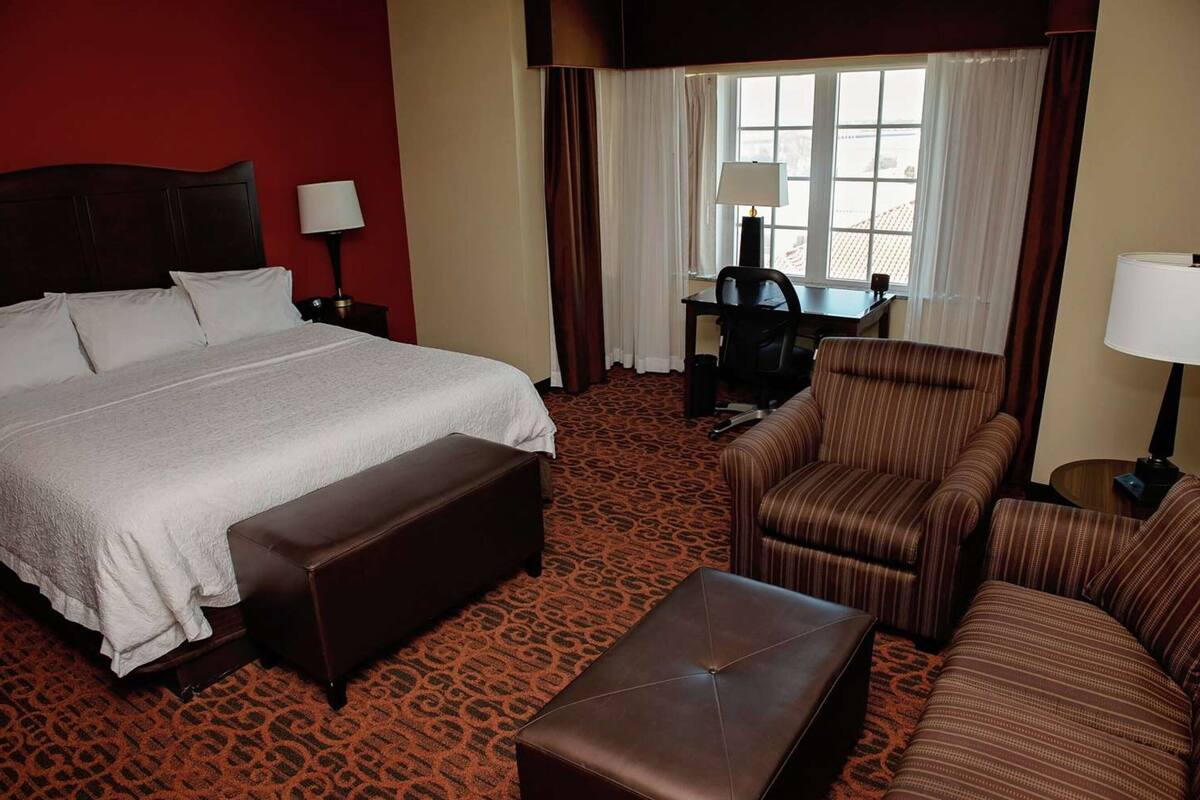 A hotel room with a large bed, a desk, two armchairs, and a small table.