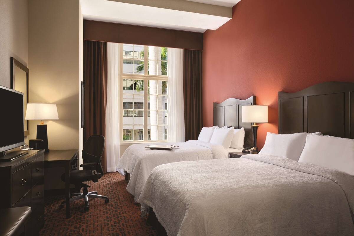 A hotel room with two beds, a desk with a lamp, a chair, and a television.