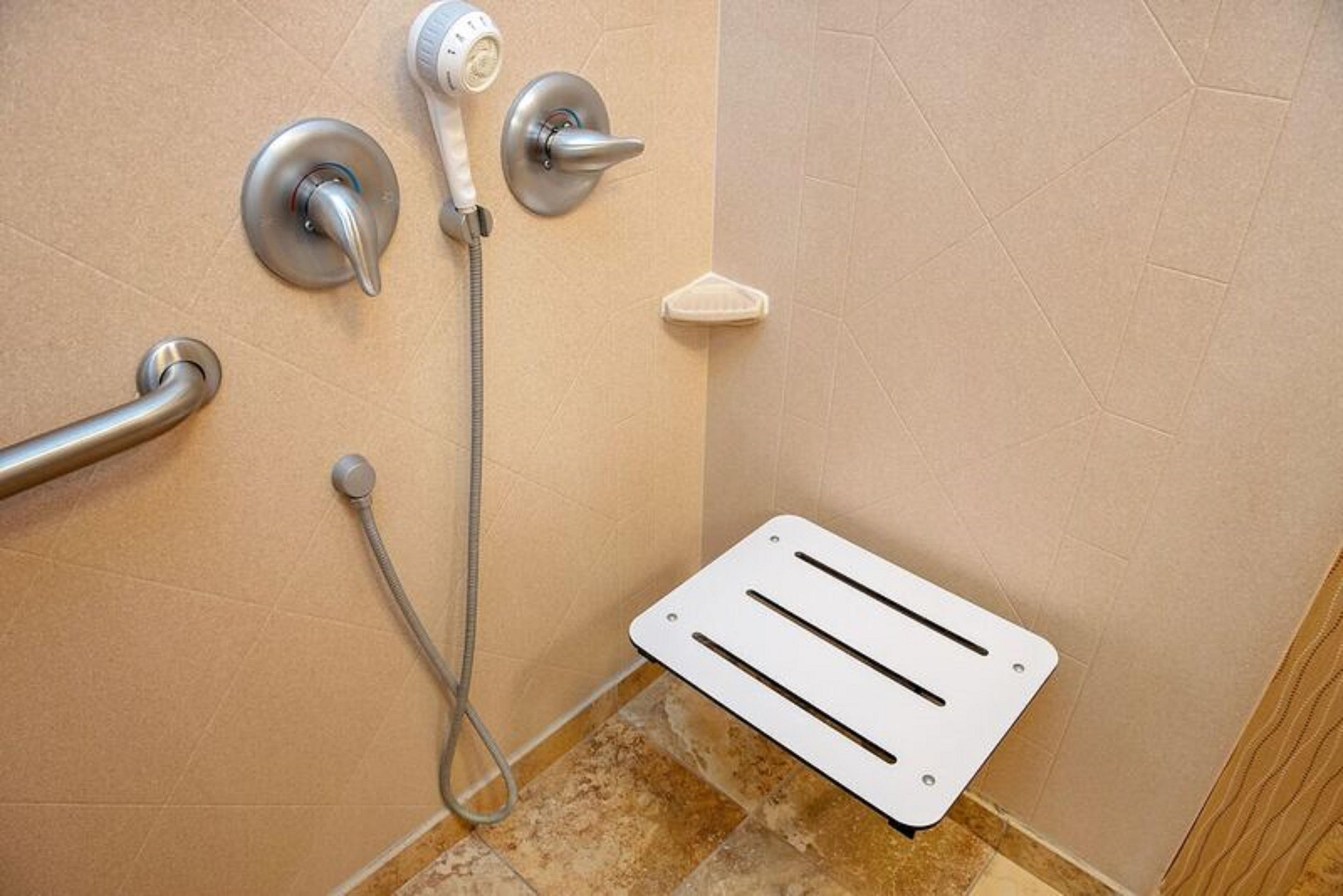 Un baño con ducha, barra de apoyo y alfombra blanca.