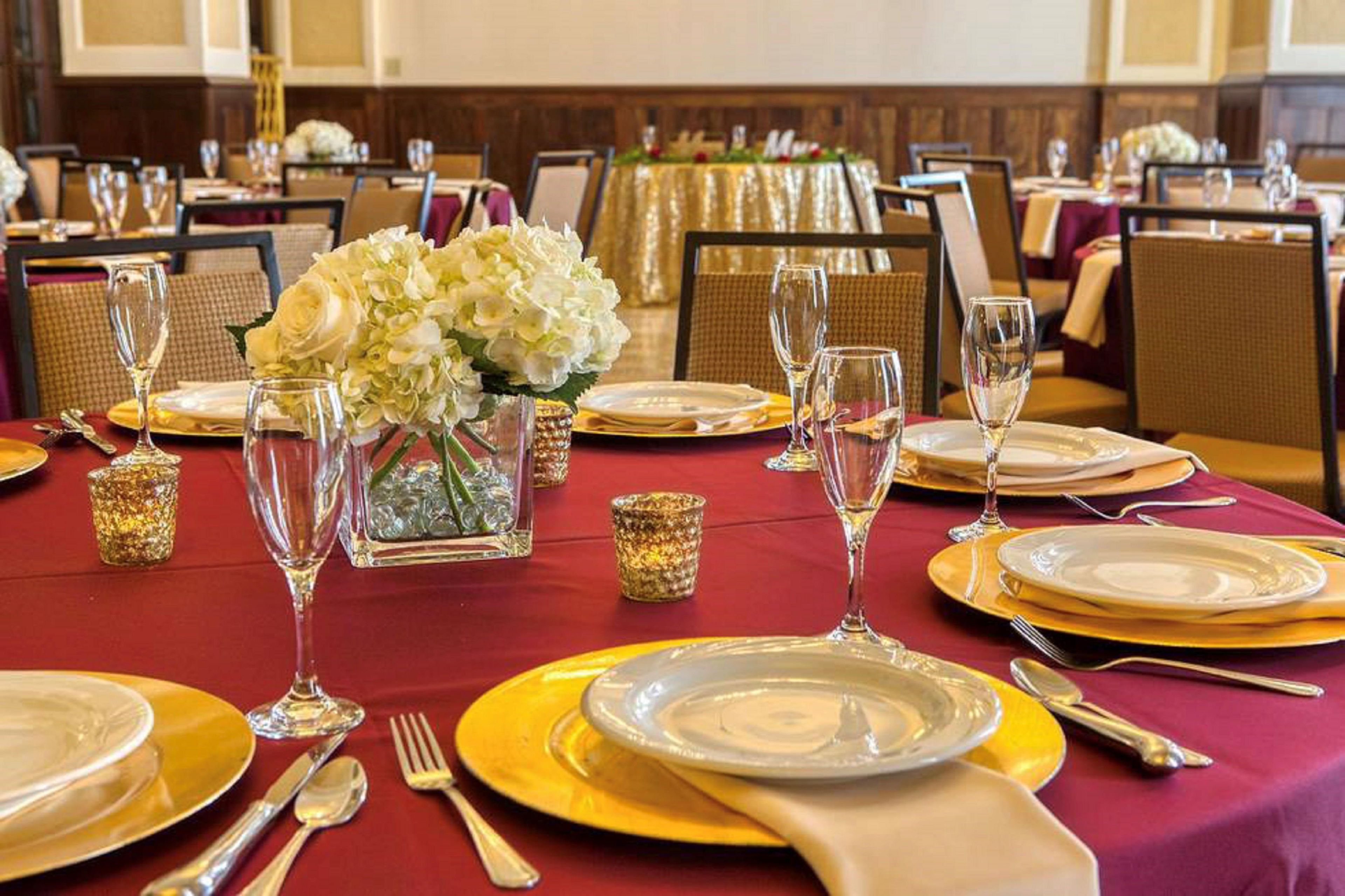 Un salón de banquetes con mesas redondas dispuestas para comer, con vajilla dorada y blanca, cristalería transparente y centros de mesa florales.