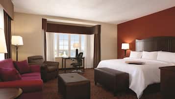A hotel room with a large bed, a desk, a chair, an ottoman, and a view of the outdoors through a window.