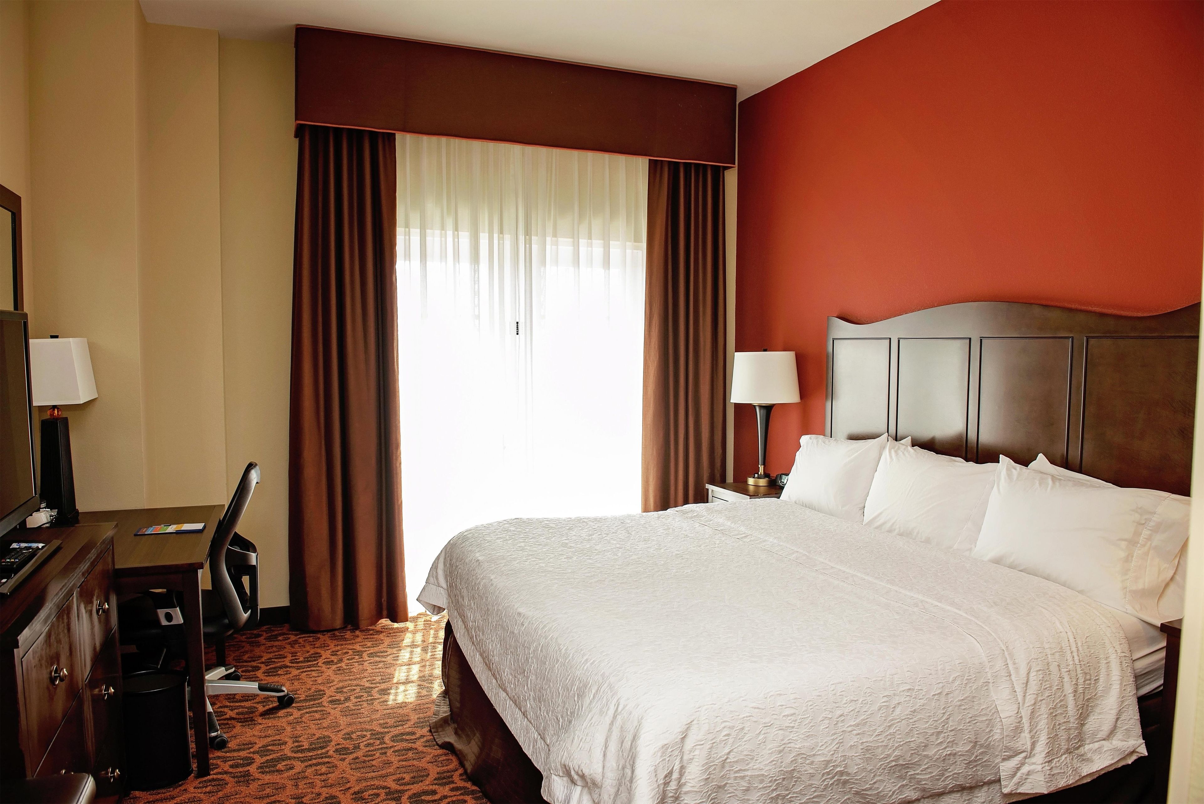 A hotel room with a bed, a desk, a chair, a lamp, and a window with curtains.