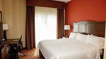 A hotel room with a bed, a desk, a chair, a lamp, and a window with curtains.