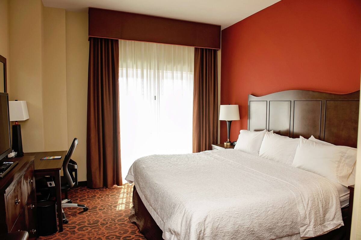 A hotel room with a large bed, a desk with a chair, a television, and a window with curtains.
