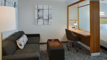 Habitación de hotel con un sofá gris oscuro, una mesita de madera, un escritorio con una silla, un espejo grande y una cama con cabecera blanca.