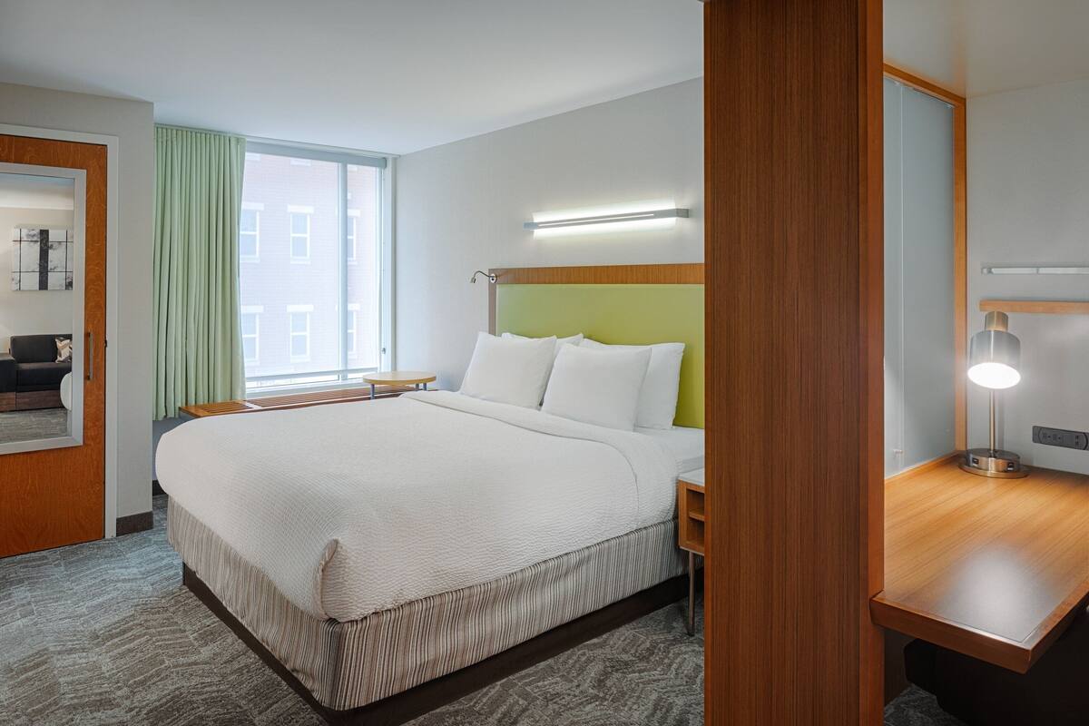 A hotel room with a large bed, a bedside table, a desk, a mirror, and a window with curtains.