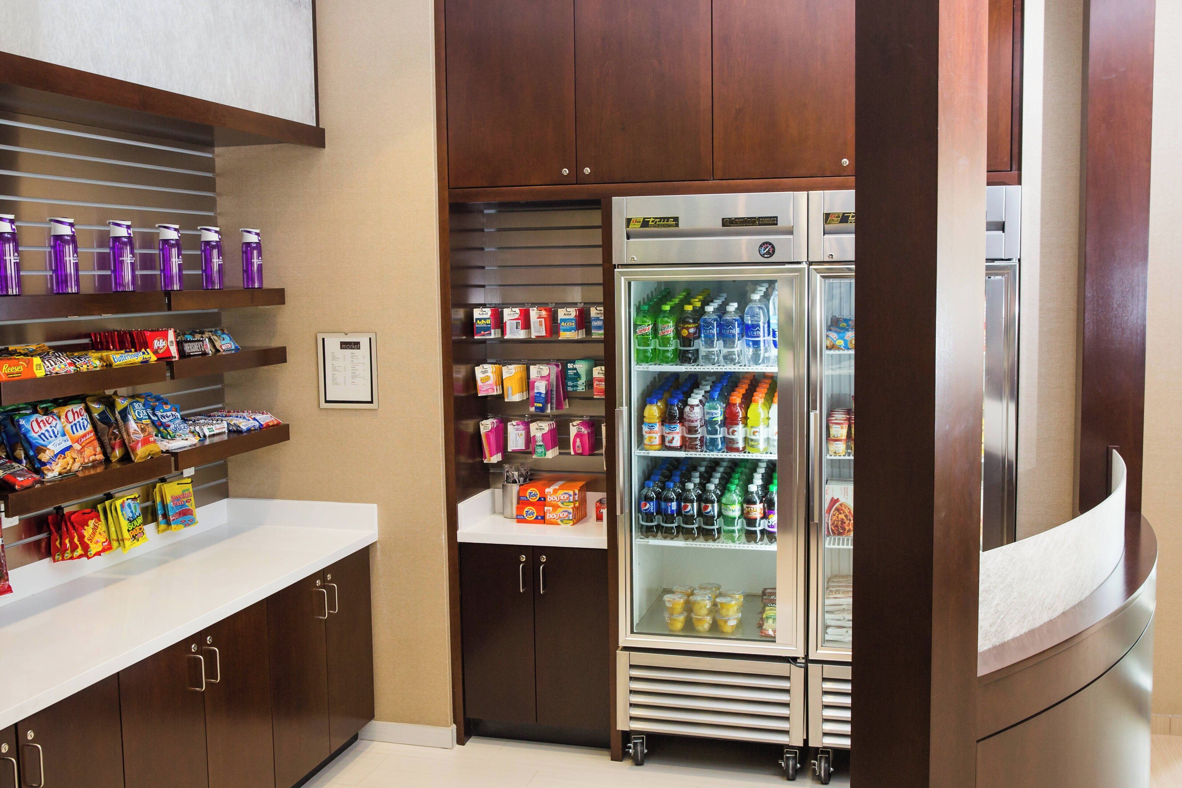Una kitchenette con un refrigerador lleno de bebidas y aperitivos, un microondas y una repisa con agua embotellada y jugo.