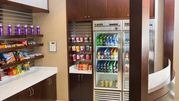 Una kitchenette con un refrigerador lleno de bebidas y aperitivos, un microondas y una repisa con agua embotellada y jugo.